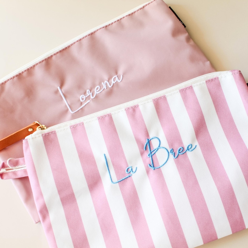 May include: Two zippered pouches. One is solid dusty pink with the name Lorena embroidered in white thread. The other is white with vertical pink stripes and the name La Bree embroidered in light blue thread. Both have white zippers.