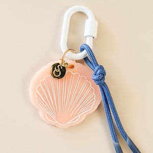 May include: A pink seashell-shaped keychain with a white outline and a gold charm. The keychain is attached to a white carabiner clip and a blue rope. The charm has a gold letter "S" on it.