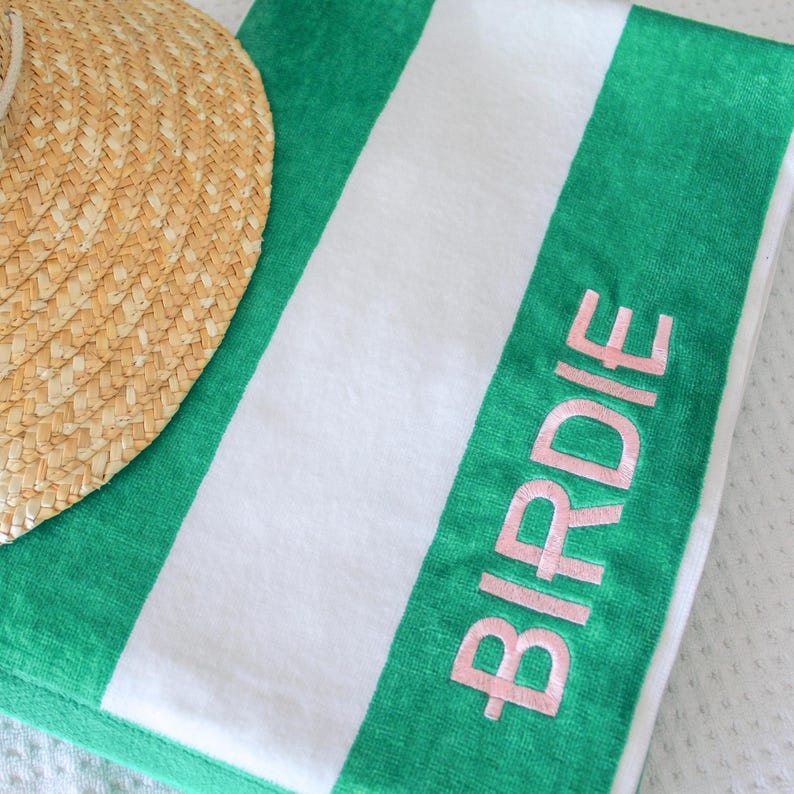 May include: A green and white striped beach towel with the name "Birdie" in pink lettering. A straw sun hat is partially visible in the upper left corner. The towel is folded and appears to be made of a soft material.