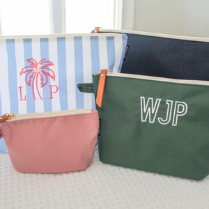May include: A collection of four zippered pouches in various colors and patterns. One pouch features blue and white vertical stripes with a pink palm tree and the letters LP. Another is solid dark green with the white letters WJP. A small pink pouch and a solid navy blue pouch are also included.