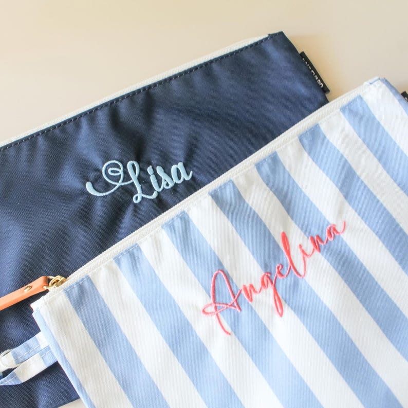 May include: Two zippered pouches, one solid navy blue with the name Lisa embroidered in light blue, and one with white and light blue vertical stripes featuring the name Angelina embroidered in coral pink thread on the front surface.