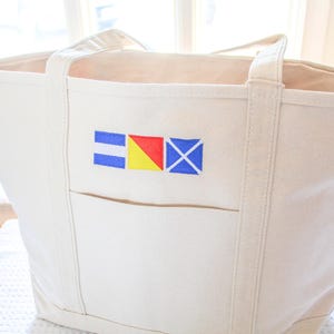 May include: Cream-colored canvas tote bag with a front pocket and sturdy handles. The bag features an embroidered nautical flag design in blue, red, and yellow. A practical and stylish accessory for carrying essentials.