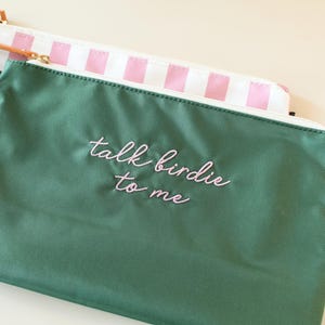 May include: A dark green zippered pouch with the embroidered text "talk birdie to me" in light pink cursive script. Behind it sits a second pouch featuring a white and light pink vertical striped pattern with a gold-toned zipper pull.