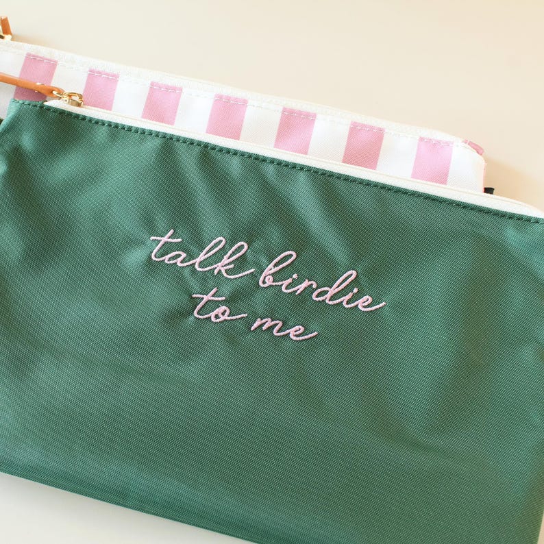 May include: A dark green zippered pouch with the embroidered text "talk birdie to me" in light pink cursive script. Behind it sits a second pouch featuring a white and light pink vertical striped pattern with a gold-toned zipper pull.