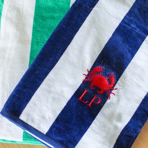 May include: A blue and white striped beach towel with a red crab embroidered on the corner. The towel also has the embroidered initials "LP" in red. A green and white striped towel is visible in the background.