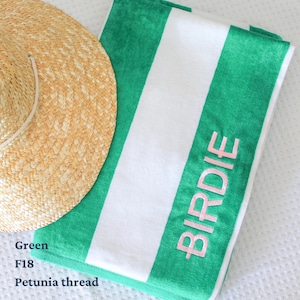 May include: A green and white striped beach towel with the name "BIRDIE" embroidered in pink. A straw sun hat rests on the corner of the towel. The towel is folded and appears soft and plush.