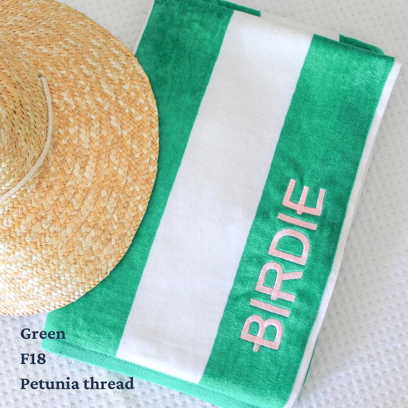 May include: A green and white striped beach towel with the name "BIRDIE" embroidered in pink. A straw sun hat rests on the corner of the towel. The towel is folded and appears soft and plush.