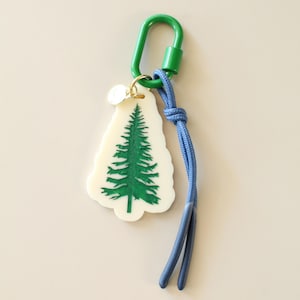 May include: A keychain featuring a cream-colored, tree-shaped charm with a green pine tree design. It includes a green carabiner clip and a blue cord. A small gold-colored charm is attached to the top of the tree charm.