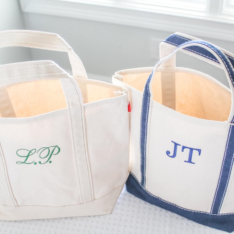 May include: Two canvas tote bags with cream-colored exteriors and handles. One bag has green embroidered initials "L.P." and the other has blue embroidered initials "J.T." The bags have a rectangular shape and are suitable for carrying items.