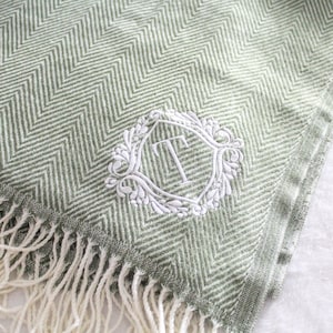 Puede incluir: Una manta verde salvia con estampado de espiga y flecos blancos. La manta presenta una letra monograma "T" bordada en blanco dentro de un marco floral decorativo. La manta est&aacute; doblada, mostrando el patr&oacute;n y los flecos.