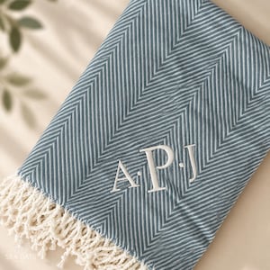 May include: A folded blue and white herringbone patterned throw blanket featuring white embroidered monogram letters A.P.J. in the center. The blanket has a decorative fringe edge made of off-white twisted cotton yarn along the bottom border.