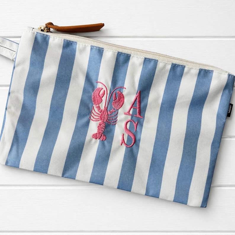May include: A rectangular zippered pouch featuring a blue and white vertical striped pattern. The center of the pouch is embroidered with a pink lobster graphic and the letters A and S in a matching pink thread. A brown leather zipper pull is attached.