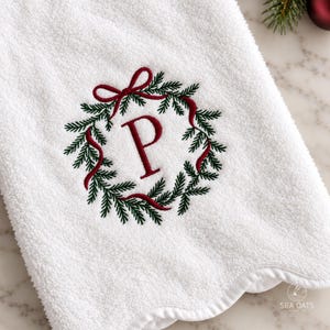 May include: A white terry cloth hand towel with a scalloped edge, featuring an embroidered design of a green pine wreath tied with a red ribbon and the letter P in the center. The text SEA OATS is visible in the bottom right corner.