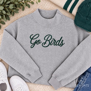May include: A heather gray long-sleeve sweatshirt with a mock neck collar. The front features the text Go Birds embroidered in dark green cursive script. The garment has ribbed cuffs and a ribbed hem, providing a structured fit.
