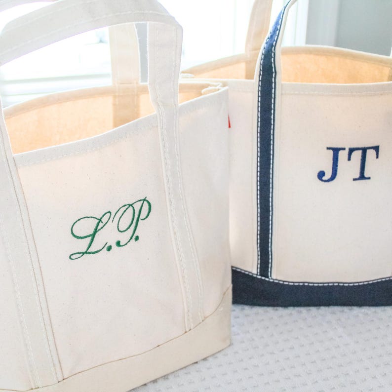 May include: Off-white canvas tote bags with embroidered initials. One bag has green initials "L.P.", the other has blue initials "J.T.". Both bags have sturdy handles and a navy blue trim.