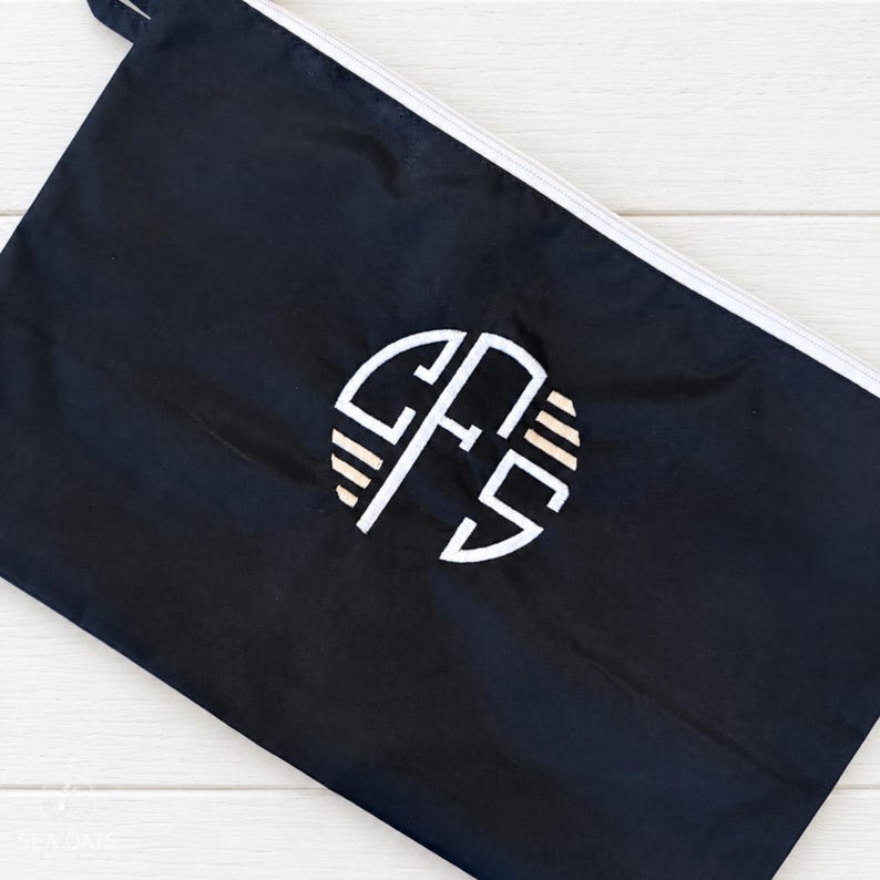 May include: A black rectangular zippered pouch featuring a white circular monogram logo in the center. The logo consists of the letters CFS stylized with horizontal lines on the sides. The pouch has a white zipper closure along the top edge.