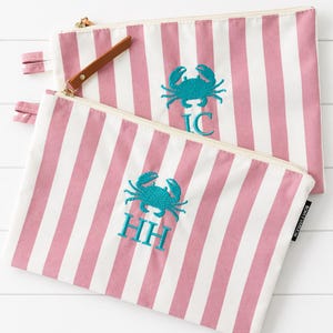 May include: Two rectangular zippered pouches featuring a vertical pink and white striped pattern. Each pouch displays an embroidered teal crab graphic centered above monogrammed letters, with one reading IC and the other reading HH in teal thread.