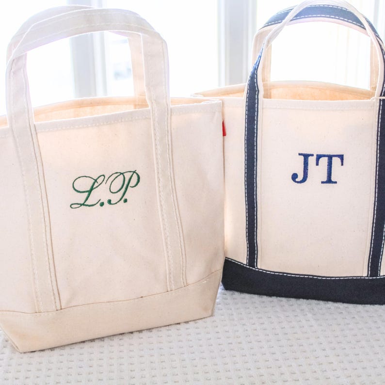 May include: Two off-white canvas tote bags with embroidered initials. One bag has green initials "L.P." and the other has blue initials "JT." Both bags have sturdy handles and a simple, classic design, perfect for everyday use.