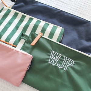 May include: A collection of four rectangular zipper pouches in various colors and patterns. One is solid navy, one features green and white vertical stripes, one is solid dark green with the white embroidered letters WJP, and one is solid dusty pink.