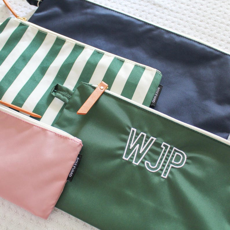 May include: A collection of four rectangular zipper pouches in various colors and patterns. One is solid navy, one features green and white vertical stripes, one is solid dark green with the white embroidered letters WJP, and one is solid dusty pink.