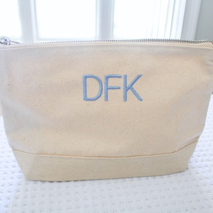 May include: Cream-colored canvas cosmetic bag with a silver zipper closure. The bag features the embroidered initials "DFK" in light blue. The bag has a rectangular base and a rounded top.