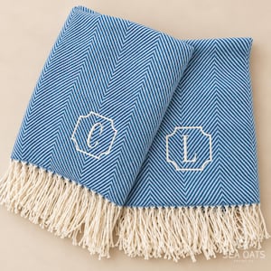 May include: Two blue and white herringbone patterned towels featuring cream-colored fringe at the bottom edges. Each towel is embroidered with a white monogrammed letter, one with a C and the other with an L, centered within a decorative frame.