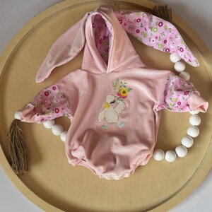 May include: A pink hooded baby onesie with bunny ears and a floral print. The onesie features an embroidered image of a duck holding flowers.