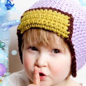 Könnte beinhalten: Ein kleines Kind trägt eine lila und gelbe Häkelmütze mit braunem Rand. Die Mütze hat ein Häkelmuster und bedeckt die Ohren des Kindes. Das Kind schaut in die Kamera und hat den Finger auf den Lippen.
