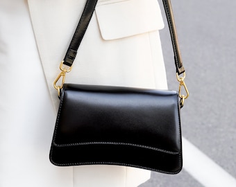 Puede incluir: Un bolso negro rectangular con una larga correa negra y herrajes dorados. El bolso está hecho de un material brillante y se lleva sobre un atuendo blanco.