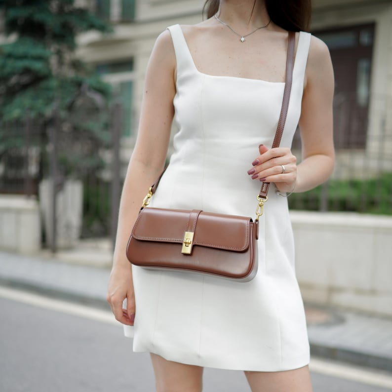 May include: A brown leather shoulder bag with a rectangular shape and a gold-colored clasp. The bag has a long, thin strap and is worn over the shoulder. The bag is a rich brown color.