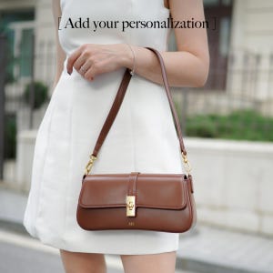 May include: A brown leather shoulder bag with a rectangular shape and a gold-colored clasp. The bag has a long, thin strap and the text "Add your personalization" is visible at the top.