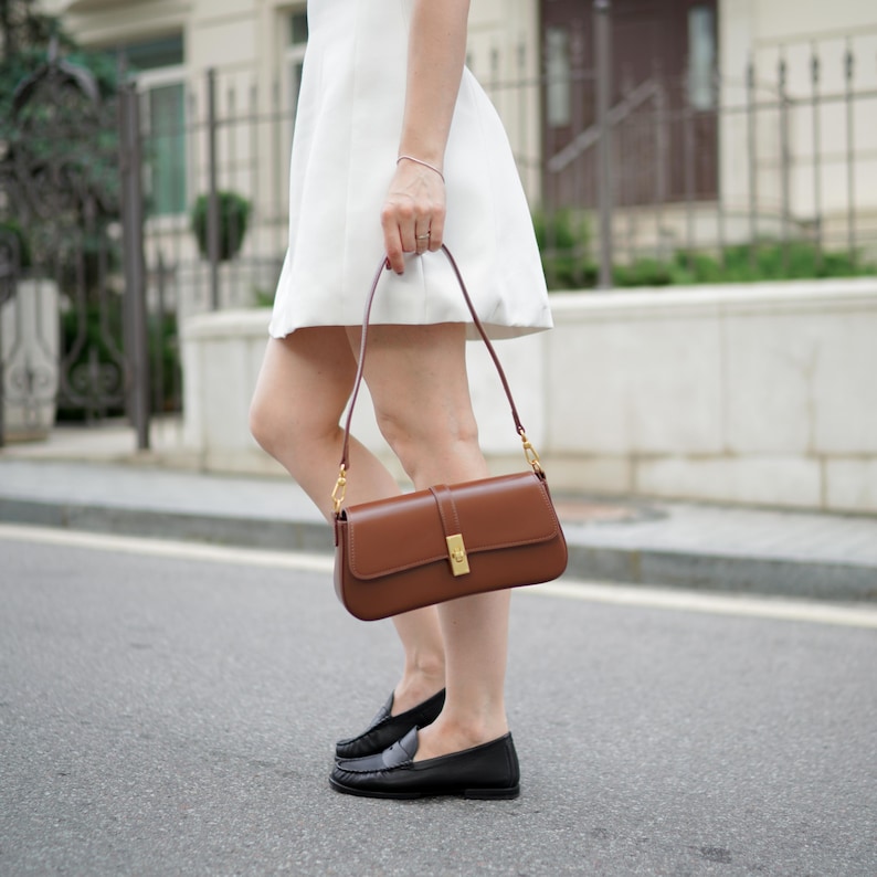 May include: A brown leather shoulder bag with a rectangular shape and a gold-colored clasp. The bag has a long, thin strap and is being held by a person wearing a white dress and black loafers.