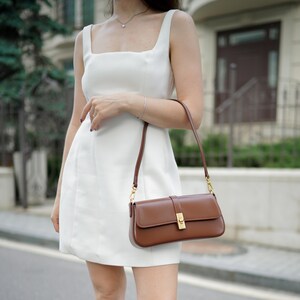 May include: A white sleeveless dress with a square neckline. A brown leather shoulder bag with a rectangular shape and a gold-colored clasp is held by the person. The bag has a long, thin strap.