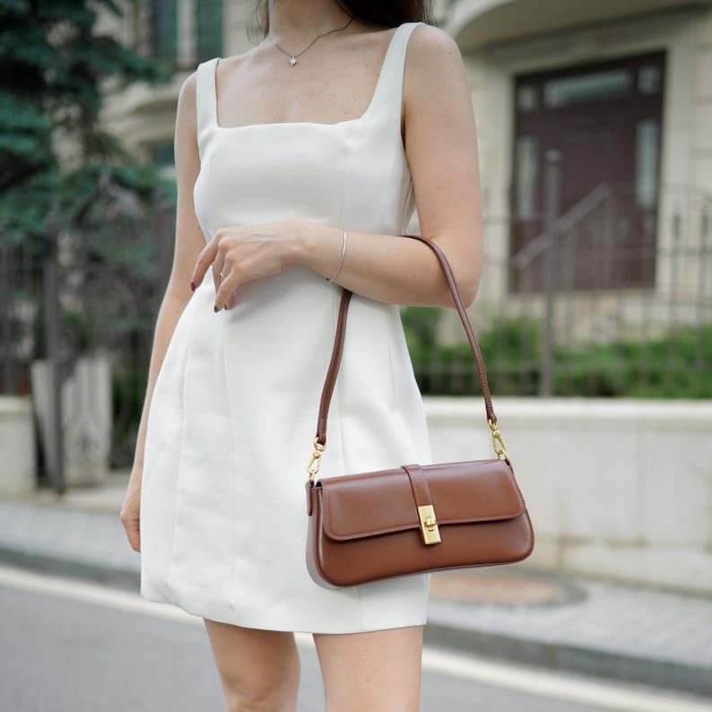 May include: A white sleeveless dress with a square neckline. A brown leather shoulder bag with a rectangular shape and a gold-colored clasp is held by the person. The bag has a long, thin strap.