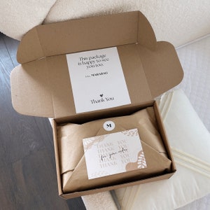 May include: An open cardboard box with a white label that reads "This package is happy to see you too." and "Thank You." Inside, the contents are wrapped in brown paper. A small card with "Thank You" is also visible.