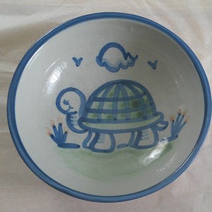 May include: A ceramic bowl with a blue rim and a whimsical turtle design. The turtle is depicted in blue and green, with a grid pattern on its shell. The bowl's interior is white, with a light green base and blue accents.