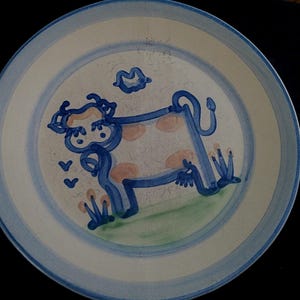 May include: A white ceramic plate with a blue and white border. The plate features a whimsical illustration of a cow in blue with pink spots, green grass, and a blue cloud. The plate has a rustic, hand-painted appearance.