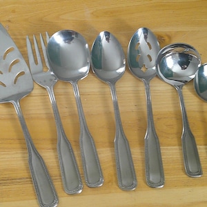 May include: A collection of stainless steel serving utensils, including a pie server, fork, spoons, and a ladle. The utensils have a simple, elegant design with a beaded edge detail. The arrangement is set against a light brown wooden surface.