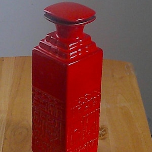 May include: A bright red ceramic bottle with a square base and a matching square stopper. The lower section of the bottle features a textured, embossed pattern on all four sides, while the upper neck transitions into a stepped, tiered design.