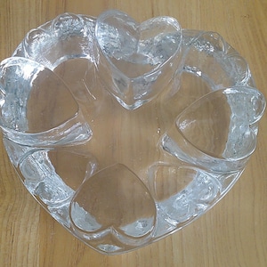 May include: A clear glass bowl featuring a circular arrangement of six molded heart shapes around the perimeter. The glass has a smooth, reflective surface with a thick, solid base and distinct heart-shaped indentations forming the outer rim.