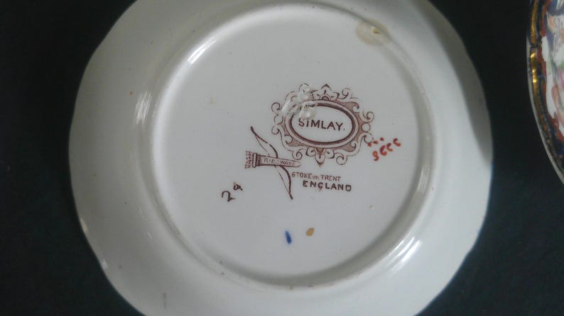 May include: A white ceramic saucer with a brown and gold design. The saucer has the words "Simlay, Stone & Trent, England" printed on the bottom.