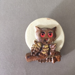 May include: A brown and yellow owl with red eyes perched on a branch in front of a white moon. The owl is a ceramic figurine.
