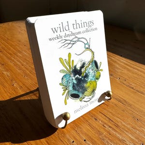 May include: A white book titled "wild things weekly daydream collection" with an illustration of aquatic plants and the author's name, "melissa moss." The book is displayed on a wooden surface, held by a brass book stand.