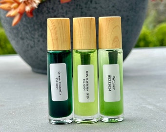 Puede incluir: Tres botellas de vidrio con rodillo y tapas de madera. Las botellas contienen líquido verde y tienen etiquetas blancas con texto. Las etiquetas dicen "German Chamomile 05232024", "100% Blueberry Seed 05232024" y "Vetiver 05232024".