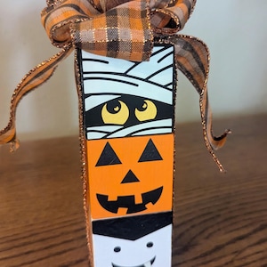 May include: A stacked Halloween-themed wooden block decoration. The top block features a mummy design, the middle block a jack-o'-lantern face, and the bottom block a vampire face. A plaid orange and brown ribbon bow adorns the top.