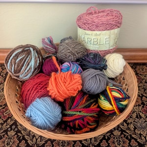 May include: A wicker basket filled with various colorful yarn balls. The yarn colors include red, orange, blue, brown, gray, and purple. A large ball of pink yarn with a label that reads "Marble" is also in the basket. The yarn is for knitting and crocheting.