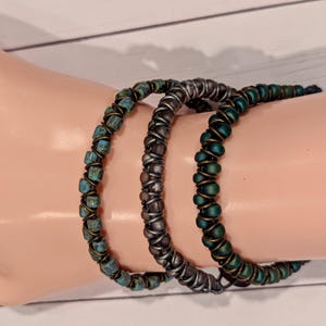 May include: Three beaded bracelets in shades of teal, silver, and green. The bracelets are wrapped with wire. The teal bracelet features square beads, while the other two have round beads. The bracelets are displayed on a light skin-toned surface.