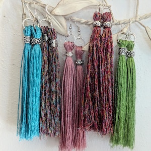 May include: A collection of tassel earrings in various colors, including turquoise, burgundy, and green. Each earring features a silver-toned hoop and decorative beadwork at the top. The tassels are long and made of a shiny, textured material, creating a bohemian aesthetic.