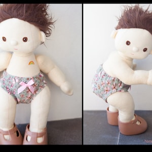 Könnte beinhalten: Eine handgemachte Stoffpuppe mit braunen Haaren, die eine Windel mit Blumenmuster und braune Schuhe trägt. Die Puppe hat einen gestickten Regenbogen auf der Brust.