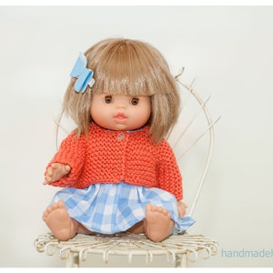 May include: A doll wearing a red knitted cardigan and a blue and white gingham dress, sitting on a white metal chair. The doll has brown eyes, light brown hair, and a blue bow in her hair.